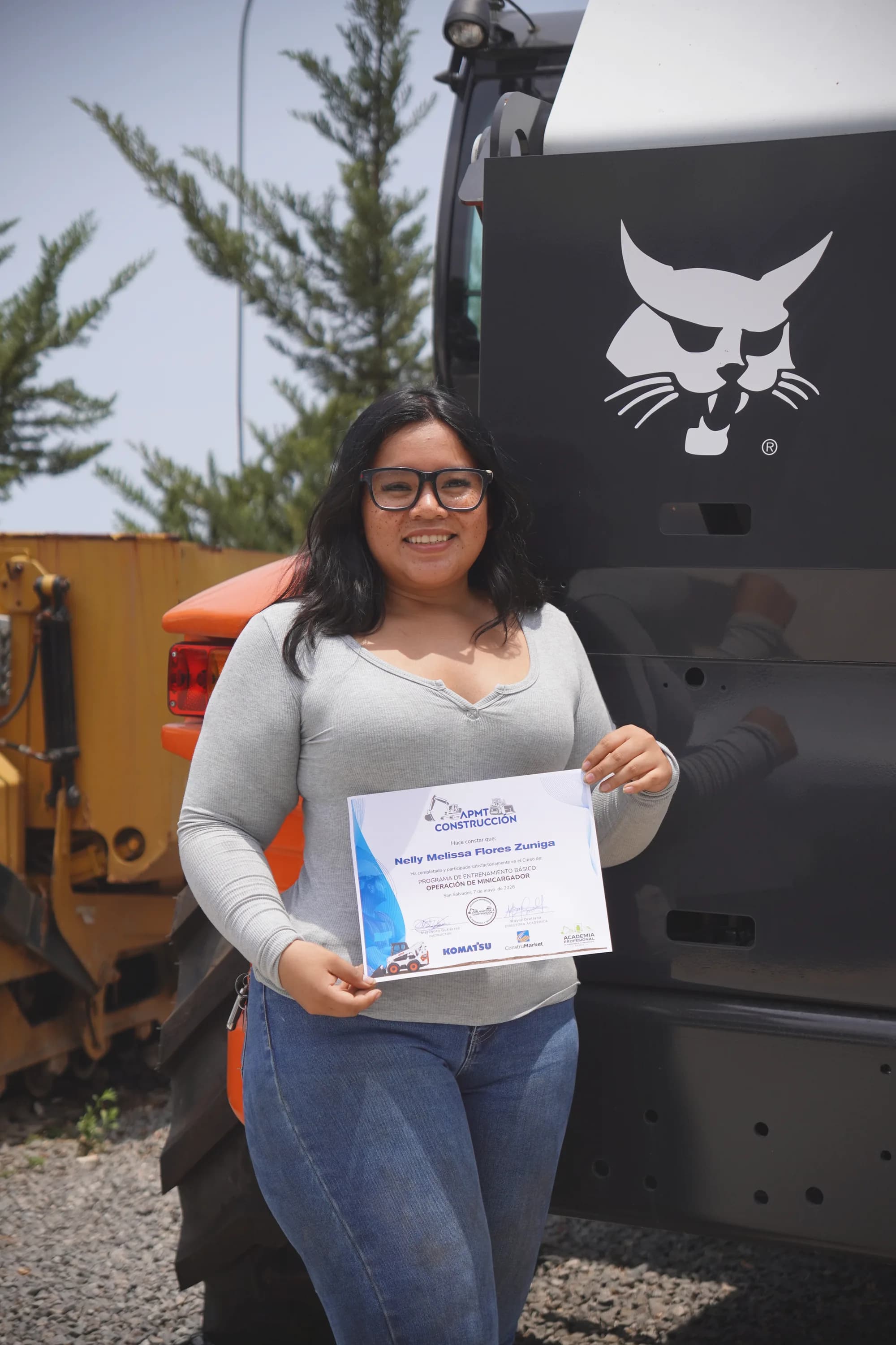 Nelly Melissa Flores Zuniga con su certificado de operación de minicargador junto a la máquina