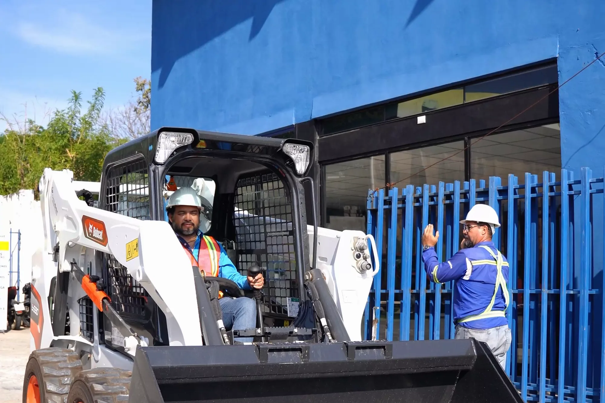 Alumno en la cabina de un minicargador con el cucharón levantado, observado por instructores